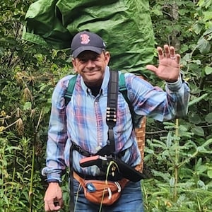 K&ouml;nnte beinhalten: Eine Person tr&auml;gt eine marineblaue Baseballkappe mit rotem B-Logo, ein lang&auml;rmeliges blau-rotes Karohemd, blaue Jeans und eine braune Bauchtasche. Auf dem R&uuml;cken wird ein gr&uuml;nes Planenb&uuml;ndel getragen und in einer Hand eine dunkle Limonadenflasche.