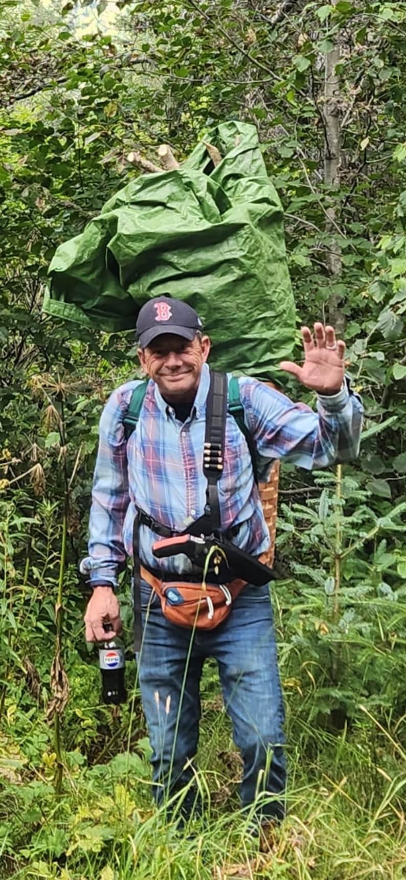 K&ouml;nnte beinhalten: Eine Person tr&auml;gt eine marineblaue Baseballkappe mit rotem B-Logo, ein lang&auml;rmeliges blau-rotes Karohemd, blaue Jeans und eine braune Bauchtasche. Auf dem R&uuml;cken wird ein gr&uuml;nes Planenb&uuml;ndel getragen und in einer Hand eine dunkle Limonadenflasche.