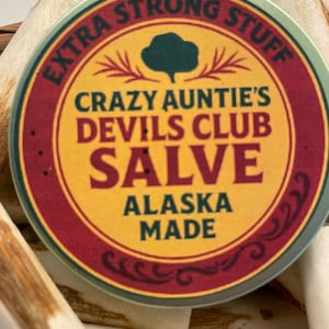 K&ouml;nnte beinhalten: Eine runde Dose mit Crazy Auntie's Devils Club Salve mit einem gelben Etikett, das dunkelgr&uuml;nen Text mit der Aufschrift Extra Strong Stuff, Crazy Auntie's Devils Club Salve und Alaska Made sowie einen dunkelroten Rand und ein gr&uuml;nes Baumsymbol zeigt.