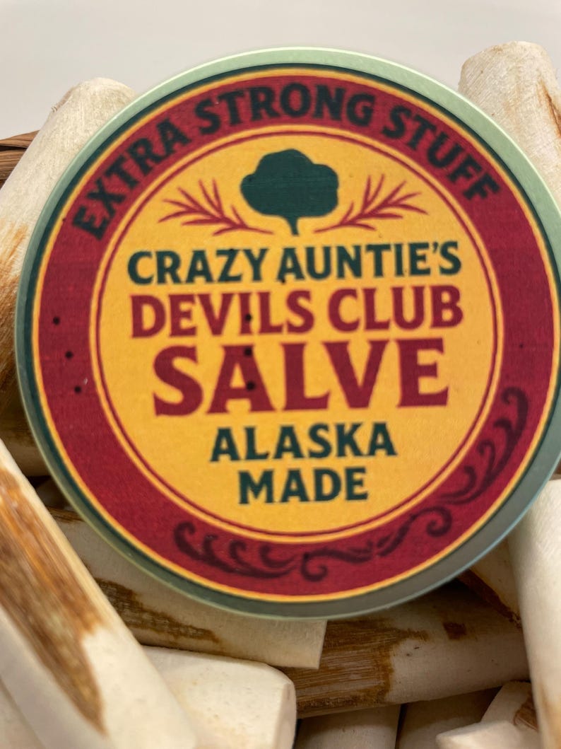 K&ouml;nnte beinhalten: Eine runde Dose mit Crazy Auntie's Devils Club Salve mit einem gelben Etikett, das dunkelgr&uuml;nen Text mit der Aufschrift Extra Strong Stuff, Crazy Auntie's Devils Club Salve und Alaska Made sowie einen dunkelroten Rand und ein gr&uuml;nes Baumsymbol zeigt.