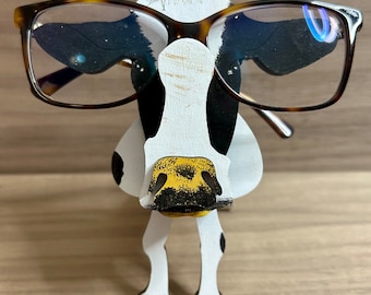 May include: A wooden cow-shaped eyeglasses holder. The holder is painted white with black spots and a yellow nose. It is holding a pair of brown-framed eyeglasses. The background is a light brown wooden surface.