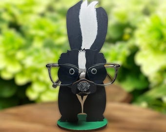 May include: A black and white wooden skunk with glasses, designed as a glasses holder. The skunk is wearing round glasses with black frames and is standing on a green base.
