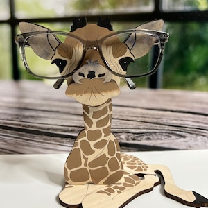 May include: A wooden giraffe-shaped eyeglasses holder with a light brown and tan spotted pattern. The giraffe is wearing clear-framed glasses. The holder is designed to sit on a flat surface and has a whimsical, decorative appeal.