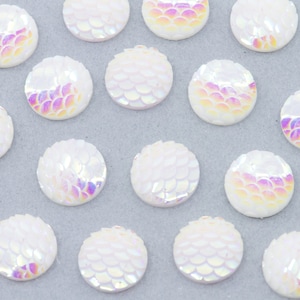 May include: A collection of white iridescent round embellishments with a scale pattern. The embellishments are scattered on a light gray surface.