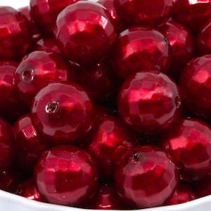 May include: A collection of faceted, round beads in a deep red hue. The beads have a glossy, reflective surface and are piled in a white bowl. These beads are suitable for jewelry making and craft projects.