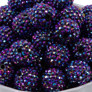 May include: A collection of large, round beads with a sparkling, iridescent finish. The beads are primarily dark purple with hints of blue, pink, and green, creating a dazzling effect. Each bead is covered in small, reflective facets. The beads are in a white bowl.