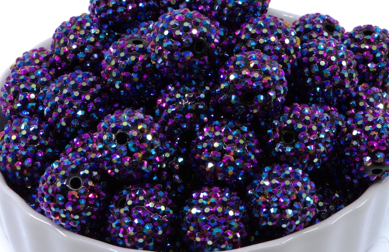 May include: A collection of large, round beads with a sparkling, iridescent finish. The beads are primarily dark purple with hints of blue, pink, and green, creating a dazzling effect. Each bead is covered in small, reflective facets. The beads are in a white bowl.