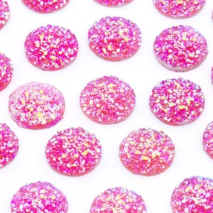 May include: A collection of round, pink, sparkly embellishments with a textured, glittery surface. The decorative elements are arranged on a white background. These are likely used for crafts or jewelry making.