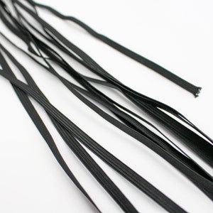 Puede incluir: Primer plano de varios cordones elásticos negros, dispuestos sobre un fondo blanco. Los cordones son finos y planos, con una textura acanalada. Los extremos de algunos cordones son visibles, mostrando la construcción tejida. Ideal para proyectos de costura y manualidades.