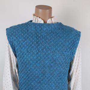 May include: A teal blue knitted vest with a textured pattern. The vest is sleeveless and worn over a white collared shirt with black polka dots. The model is wearing blue jeans.