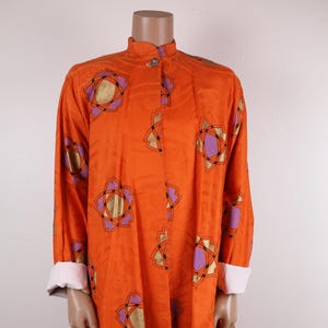 May include: An orange, long-sleeved dress with a mandarin collar and a matching skirt. The dress features a repeating floral pattern in gold, purple, and black. The cuffs are white. The outfit is displayed on a mannequin.