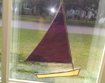 May include: A stained glass sailboat hanging in a window. The sail is a deep purple colour and the hull is a light yellow.