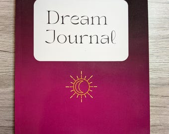 May include: A Dream Journal notebook with a gradient burgundy and black cover. The notebook features the text "Dream Journal" in a white rectangle, and a gold sun and crescent moon design.
