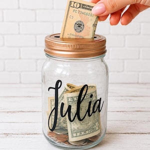 May include: A clear glass jar with a metallic copper-colored lid featuring a coin slot. The front of the jar displays the name Julia in black cursive script. Inside the jar are several rolled-up US dollar bills and various metal coins.