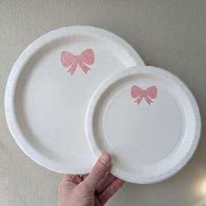 May include: Two white paper plates, one large and one small, each featuring a pink glitter bow design. The plates are held by a hand, highlighting the contrast between the white surface and the delicate pink bows. A simple, elegant design.