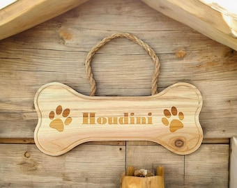 Könnte beinhalten: Ein hölzernes Hundesknochen-Schild mit dem eingravierten Namen "Houdini". Das Schild hat auf beiden Seiten des Namens zwei Pfotenabdrücke.