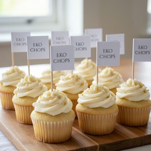 May include: A dozen cupcakes with white frosting and gold sprinkles are arranged on a wooden board. Each cupcake has a small flag that reads "EKO CHOPS". The cupcakes are a light yellow color.