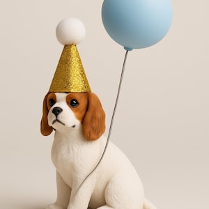 May include: A white and brown Cavalier King Charles Spaniel puppy wearing a gold glitter party hat with a white pom-pom on top. The puppy is sitting and holding a blue balloon.