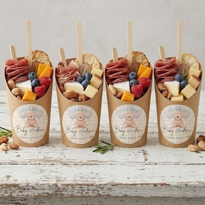 May include: Four paper cups filled with charcuterie, cheese, berries, and nuts, each topped with a wooden skewer and a cracker. Each cup has a sticker that says "IT'S A BOY!" and "Baby Andrews COMING AUGUST 2024".