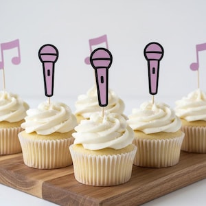 Pode incluir: Seis cupcakes com cobertura branca, cada um coberto com um palito rosa de microfone ou nota musical. Os cupcakes estão em uma tábua de madeira, perfeitos para uma festa com tema musical.