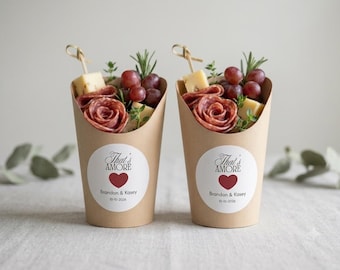 May include: Two brown paper cones filled with salami roses, cubed cheese, red grapes, and sprigs of rosemary. Each cone features a white circular sticker with the text That's AMORE, a red heart, Brandon & Kasey, and the date 10-10-2026.