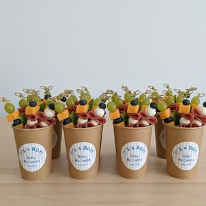 May include: Several paper cups filled with appetizers, including grapes, cheese, and meat, topped with skewers. Each cup has a sticker that reads "IT'S A BOY!" and "Baby McCombs". The cups are arranged on a light brown surface.