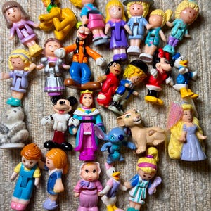 May include: A collection of small plastic figurines featuring various cartoon characters. The figurines are in various colors and depict characters such as Mickey Mouse, Donald Duck, and Disney princesses. The toys are arranged on a textured surface.