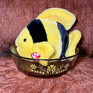 May include: A plush yellow and black fish toy with a red tag, resting inside a clear, round, glass dish. The dish has a floral design in gold. The background is a fuzzy, brown surface.