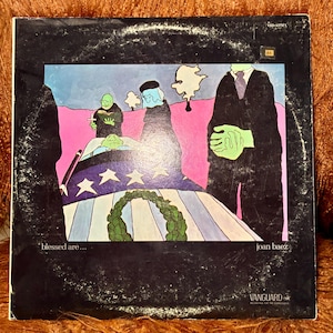 May include: Vintage vinyl record album cover for Joan Baez's "Blessed Are..." featuring an illustration. The artwork depicts figures in black suits and green skin, with a coffin draped in an American flag. The album cover has a black background with the Vanguard Records logo.