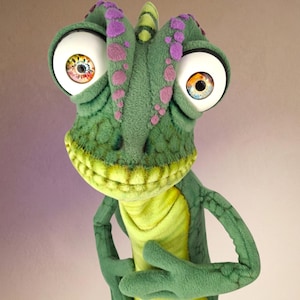 May include: A plush chameleon toy featuring a textured green body with purple spots along the head and back. The character has large, round white eyes with detailed multicoloured irises, a wide yellow mouth, and a light yellow chest panel.