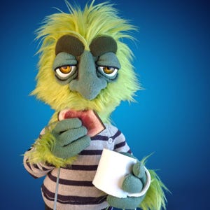 May include: A puppet with shaggy lime green fur, dark green fleece facial features, and amber eyes. It wears a grey and dark blue striped long-sleeve shirt. The puppet holds a piece of toast with red jam in one hand and a white mug in the other.