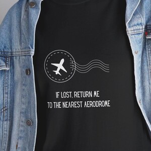 May include: Black t-shirt featuring a white airplane graphic within a circle, accompanied by three curved lines. The text reads "IF LOST, RETURN ME TO THE NEAREST AERODROME". The person is wearing a denim jacket.