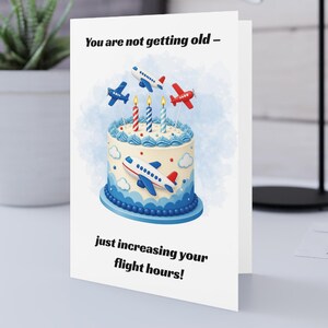 May include: A white greeting card featuring a birthday cake illustration with blue frosting, three lit candles, and small toy aeroplanes. The text reads: You are not getting old – just increasing your flight hours! The design includes clouds and blue accents.