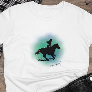 Peut inclure: T-shirt blanc avec une silhouette noire d'une personne à cheval, avec l'inscription "free spirit" en écriture manuscrite. Le motif présente un effet aquarelle bleu et vert. Le t-shirt est associé à un jean noir et des baskets.