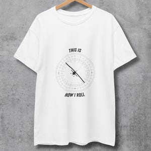 May include: White t-shirt with the text "THIS IS HOW I ROLL" and a compass-like graphic in the centre. The graphic features a circular design with a black line and a small airplane icon. The shirt is hanging on a wooden hanger.
