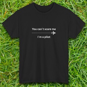 May include: Black t-shirt with white text: "You can't scare me, I'm a pilot." The text is centered, with a dotted line and airplane graphic separating the phrases. The shirt is displayed on a green grassy background.