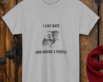 Puede incluir: Camiseta gris claro con el texto "I LIKE BATS AND MAYBE 3 PEOPLE" y un gráfico de un murciélago en blanco y negro. La camiseta es de manga corta.
