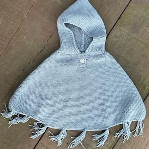 Può includere: Un poncho con cappuccio color azzurro dalla consistenza lavorata a maglia, dotato di un singolo bottone circolare sullo scollo e un orlo rifinito con nappe a frangia pendenti lungo il bordo inferiore dell'indumento.