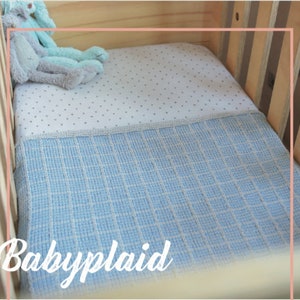 Puede incluir: Una manta para bebé azul y blanca con un patrón a cuadros se coloca sobre una sábana de cuna blanca con pequeños puntos grises. La manta está etiquetada como "Babyplaid".