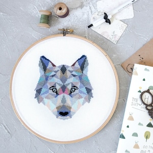 May include: A geometric wolf cross-stitch pattern in shades of blue, grey, and pink, framed in a wooden hoop.