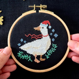 May include: A white goose wearing a red Santa hat and a red scarf is embroidered on a black fabric background in a wooden embroidery hoop. The goose is surrounded by green leaves and blue snowflakes.