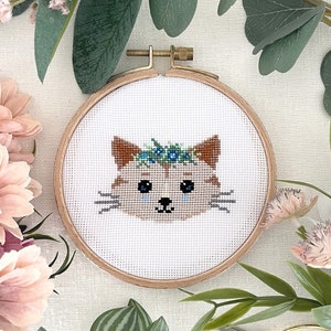 May include: A cross-stitch embroidery hoop with a cute cartoon cat wearing a flower crown. The cat is brown with blue eyes and a pink nose. The hoop is made of wood and has a white fabric background.