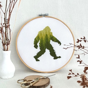 May include: A cross-stitch embroidery hoop with a green and yellow silhouette of Bigfoot walking. The hoop is on a white surface with brown branches and a pair of gold scissors.