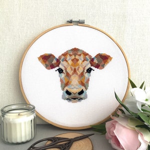 May include: A colourful cross-stitch pattern of a cow's head in a wooden embroidery hoop. The cow has brown, orange, yellow, and blue markings.