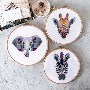 Peut inclure: Trois cerceaux de broderie avec des motifs d'animaux géométriques. Les cerceaux sont blancs avec un cadre en bois. Les motifs sont une girafe, un éléphant et un zèbre. Les animaux sont tous dans un style géométrique coloré.