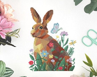 May include: A cross-stitch design of a brown rabbit surrounded by colorful flowers and greenery. A blue butterfly is near the rabbit's ear. The artwork is set against a white background, with floral and greenery accents around the edges.