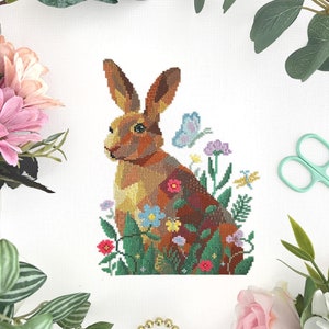 May include: A cross-stitch design of a brown rabbit surrounded by colorful flowers and greenery. A blue butterfly is near the rabbit's ear. The artwork is set against a white background, with floral and greenery accents around the edges.