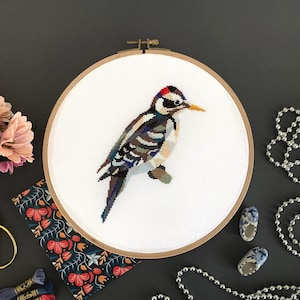 May include: A white embroidery hoop with a colorful cross-stitch design of a woodpecker. The hoop is on a gray surface with a string of silver beads.