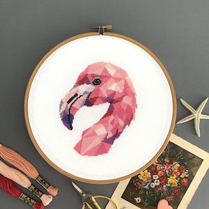 May include: A pink flamingo cross stitch pattern in a wooden embroidery hoop. The hoop is on a grey surface with pink embroidery floss, a pair of gold scissors, a white starfish, and a vintage postcard with flowers.