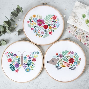 May include: Three embroidery hoops with floral designs. The first hoop features a heart-shaped floral design, the second hoop features a butterfly with floral wings, and the third hoop features a hedgehog with floral accents.